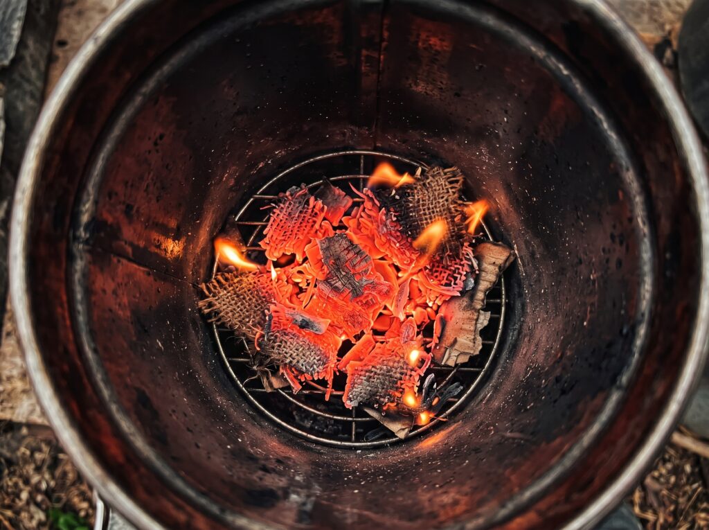 Top-down view of a glowing red bed of hot coals at the bottom of an open metal bee smoker canister, demonstrating the starter fuel layer.